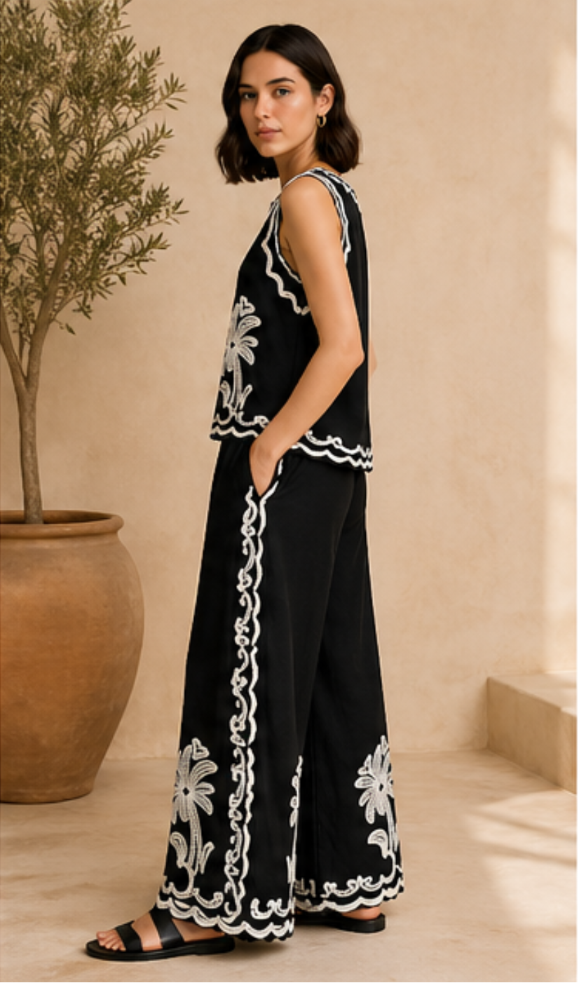 Woman wearing a black outfit with white floral embroidery in a room with a plant.