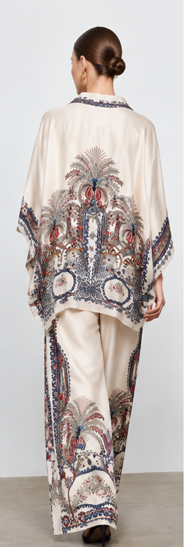 A model is shown in multiple views wearing a matching two-piece outfit consisting of a loose button-front top and wide-leg pants, both in a warm off-white base with a detailed multicolor paisley and floral print. The print features navy blue, rust red, and beige tones.
