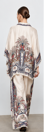 A model is shown in multiple views wearing a matching two-piece outfit consisting of a loose button-front top and wide-leg pants, both in a warm off-white base with a detailed multicolor paisley and floral print. The print features navy blue, rust red, and beige tones.