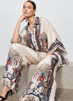 A model is shown in multiple views wearing a matching two-piece outfit consisting of a loose button-front top and wide-leg pants, both in a warm off-white base with a detailed multicolor paisley and floral print. The print features navy blue, rust red, and beige tones.